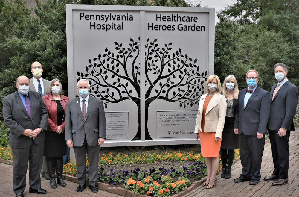 Theresa Larivee, on right, stands next to eight members of the Executive Leadership team in front of a large gray sign with the image of a tree, labeled Healthcare Heroes Garden, at Pennsylvania Hospital